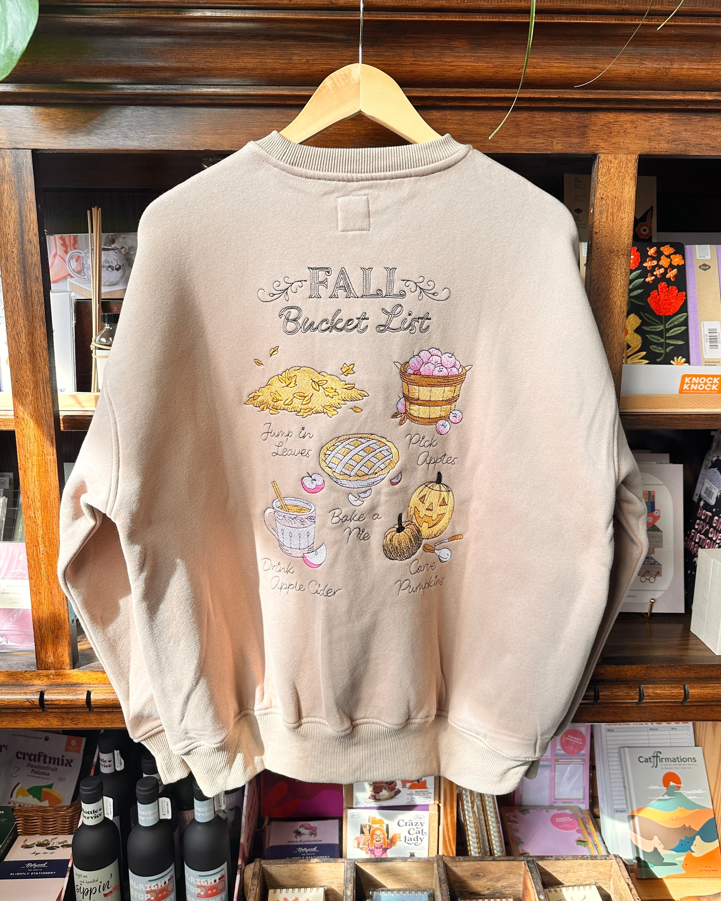 A light beige crewneck sweatshirt with a 'Fall Bucket List' embroidered print.