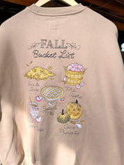 A light beige crewneck sweatshirt. The sweatshirt features a graphic print titled 'FALL Bucket List' with illustrations of autumn treats like pumpkin pie, caramel apple pie, chocolate chip cookie, and pumpkin.