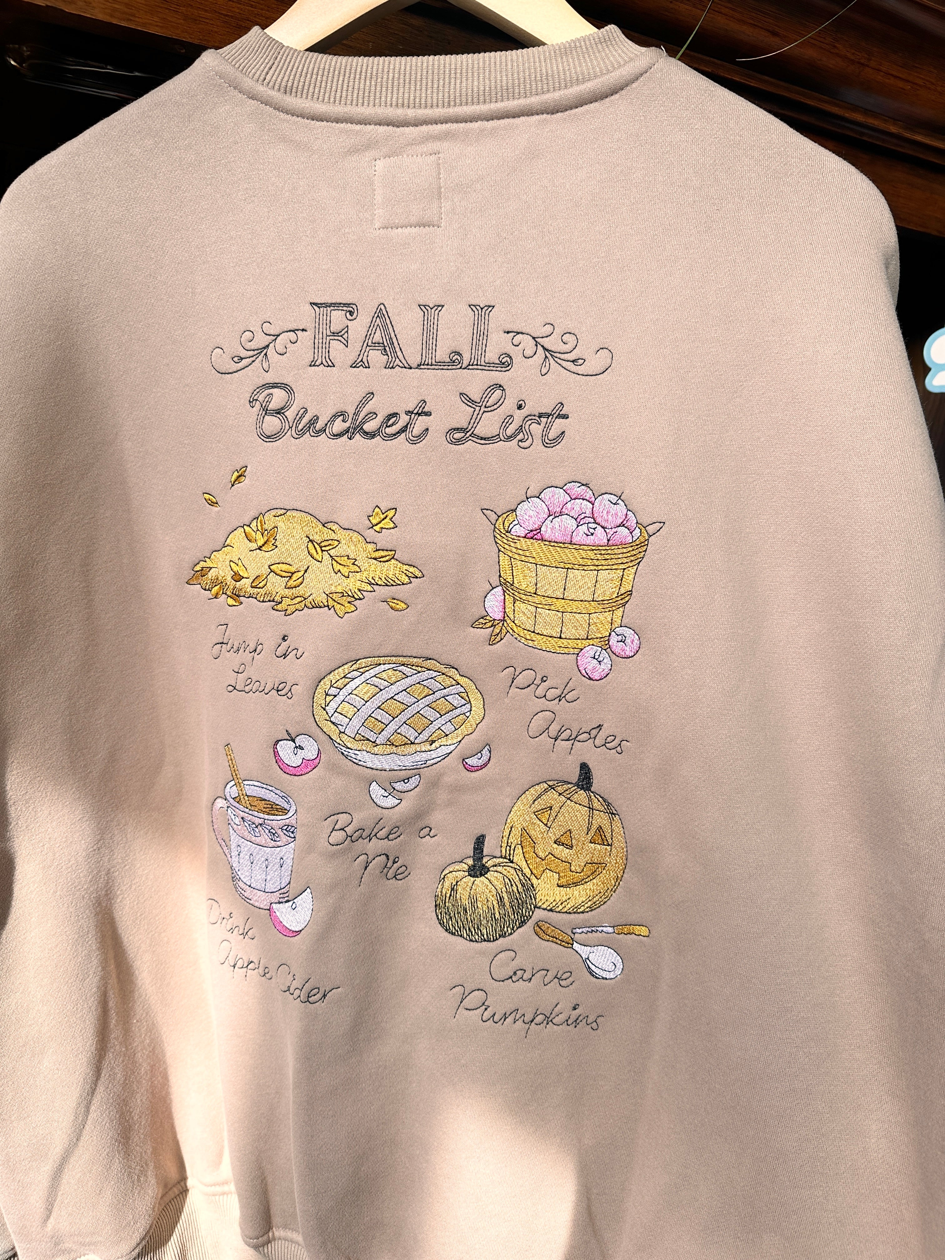 A light beige crewneck sweatshirt. The sweatshirt features a graphic print titled 'FALL Bucket List' with illustrations of autumn treats like pumpkin pie, caramel apple pie, chocolate chip cookie, and pumpkin.