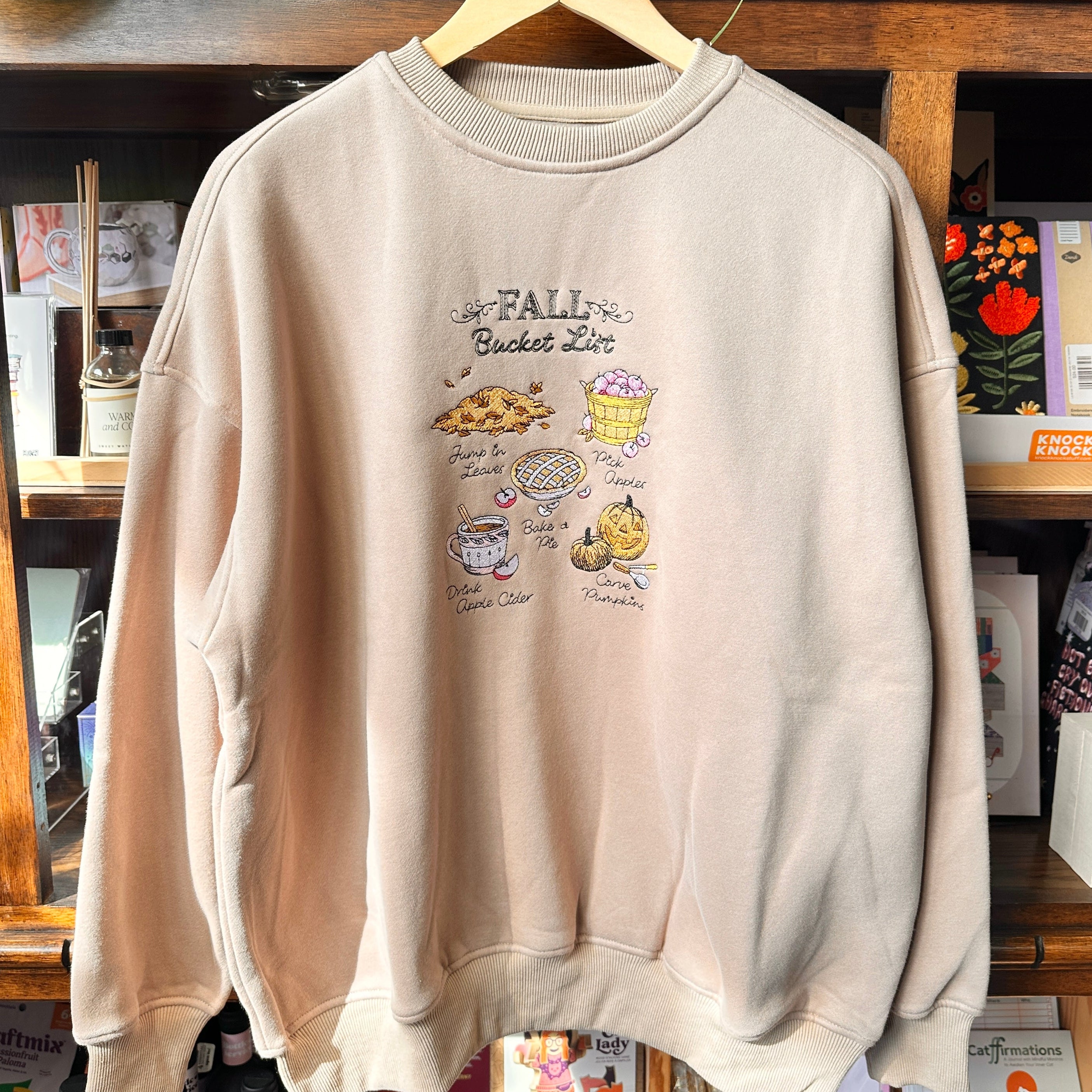 A light beige crewneck sweatshirt with a 'Fall Bucket List' embroidered print.