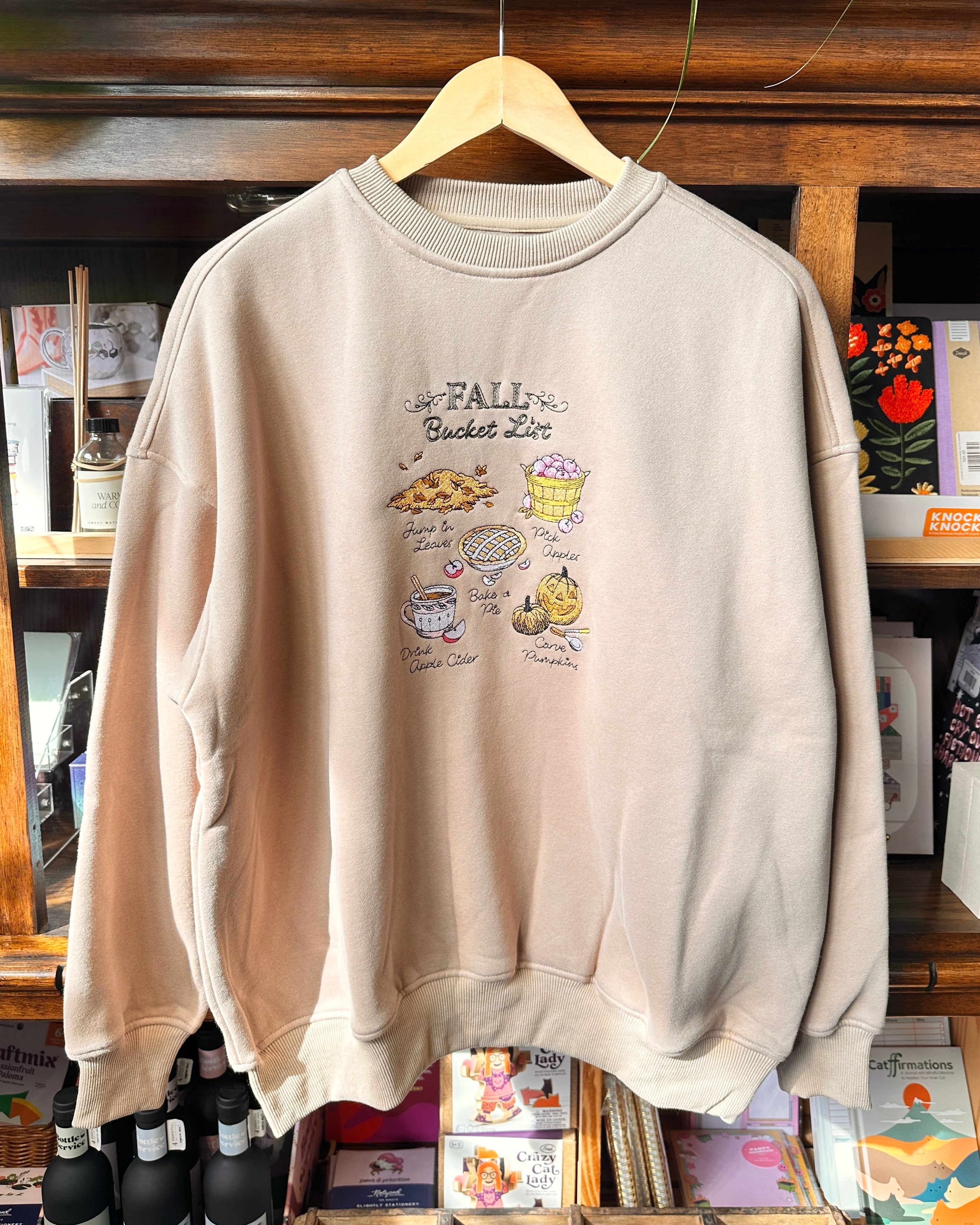 A light beige crewneck sweatshirt with a 'Fall Bucket List' embroidered print.