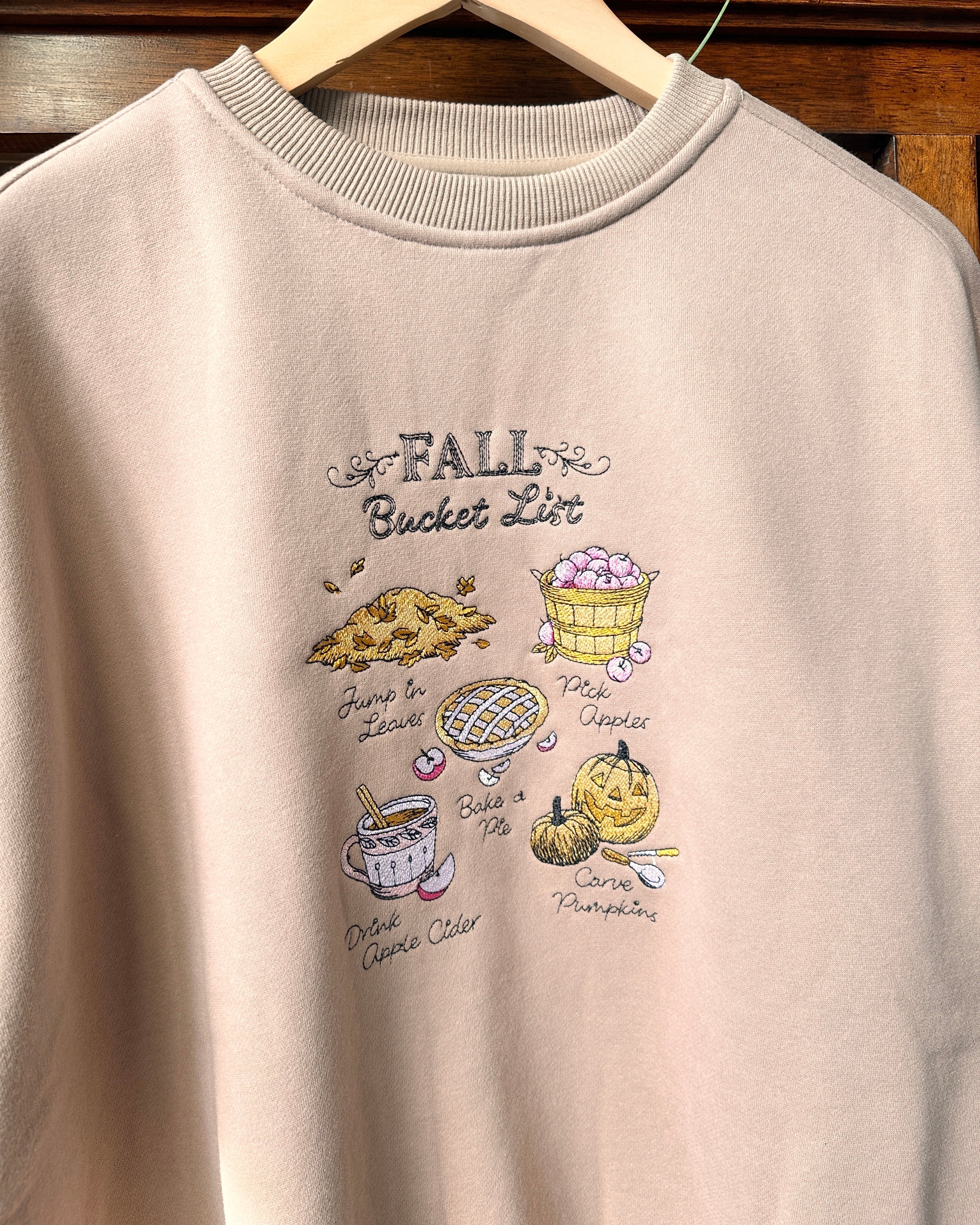 A light beige crewneck sweatshirt. The sweatshirt features a graphic print titled 'FALL Bucket List' with illustrations of autumn treats like pumpkin pie, caramel apple pie, chocolate chip cookie, and pumpkin.