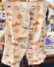 A sleeveless cream-colored vest with an all-over autumn print featuring pumpkins, waffle patterns, cozy sweaters, and fall leaves.