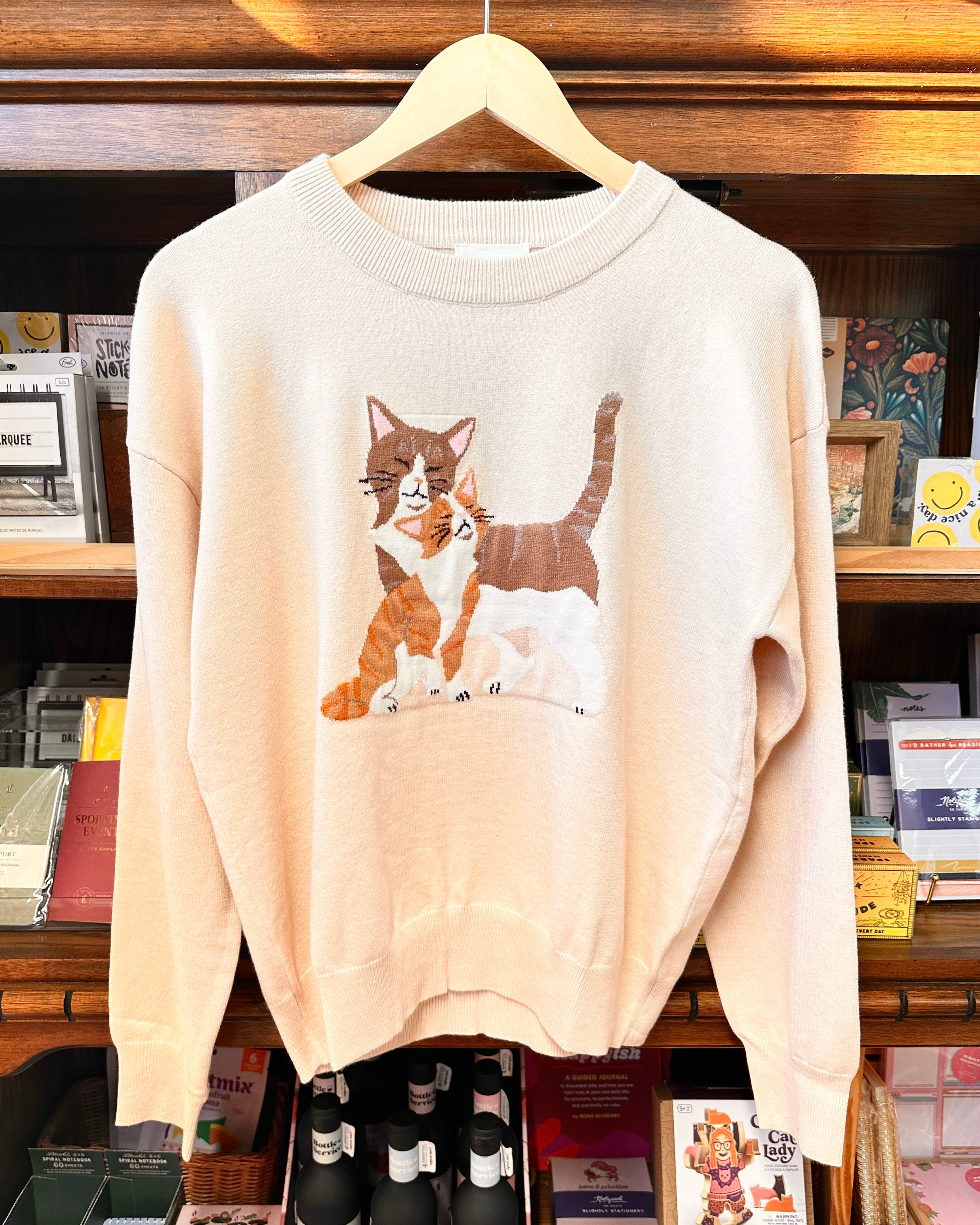 A beige crewneck sweater featuring a graphic illustration of an orange and white cat as well as a brown and white cat.