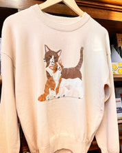 A beige crewneck sweater featuring a graphic illustration of an orange and white cat as well as a brown and white cat.