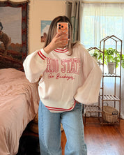 A woman takes a mirror selfie in a bedroom, wearing a cream-colored bubble-sleeve sweatshirt with red and white lettering that reads 'OHIO STATE go buckeyes', paired with light blue jeans.