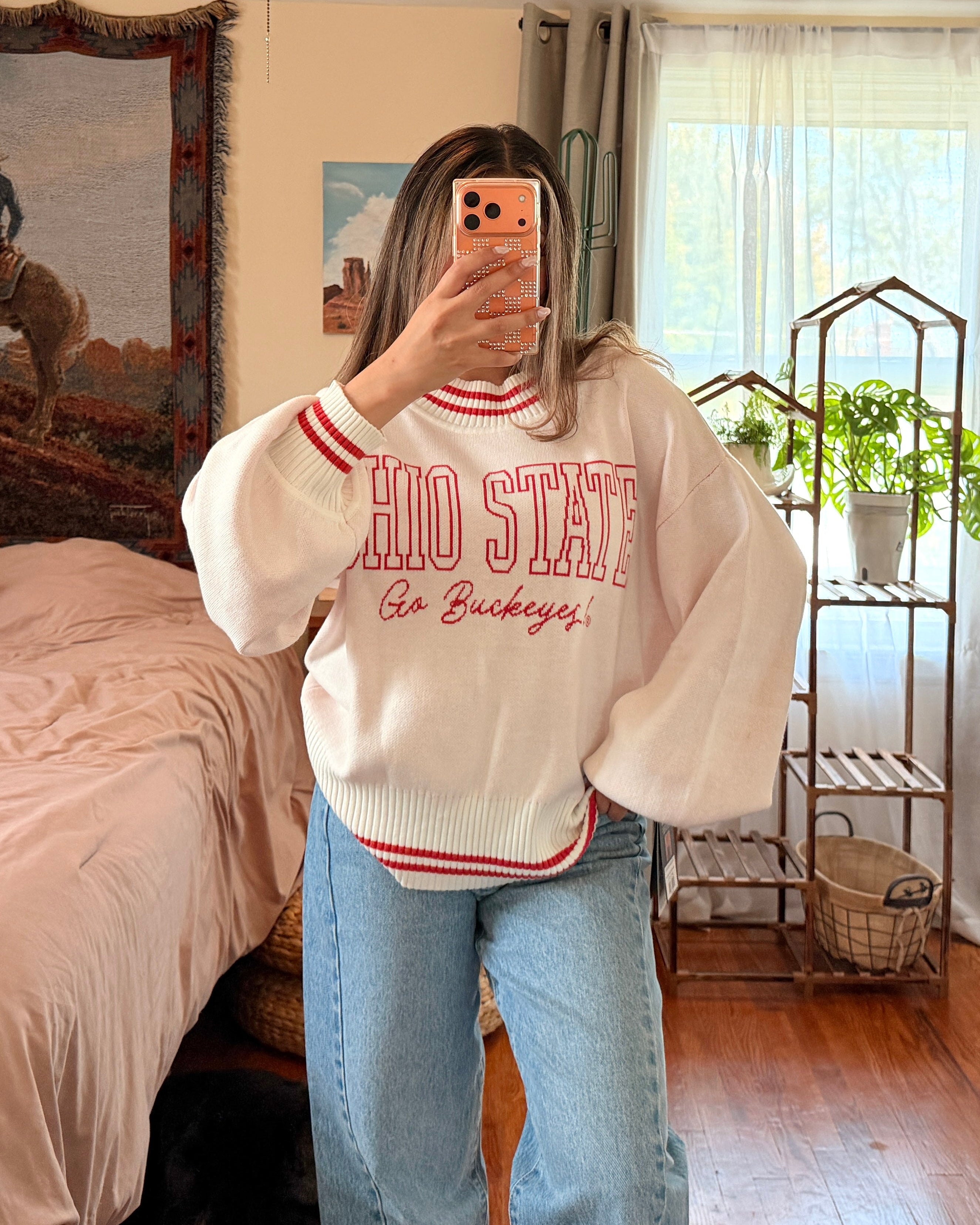 A woman takes a mirror selfie in a bedroom, wearing a cream-colored bubble-sleeve sweatshirt with red and white lettering that reads 'OHIO STATE go buckeyes', paired with light blue jeans.