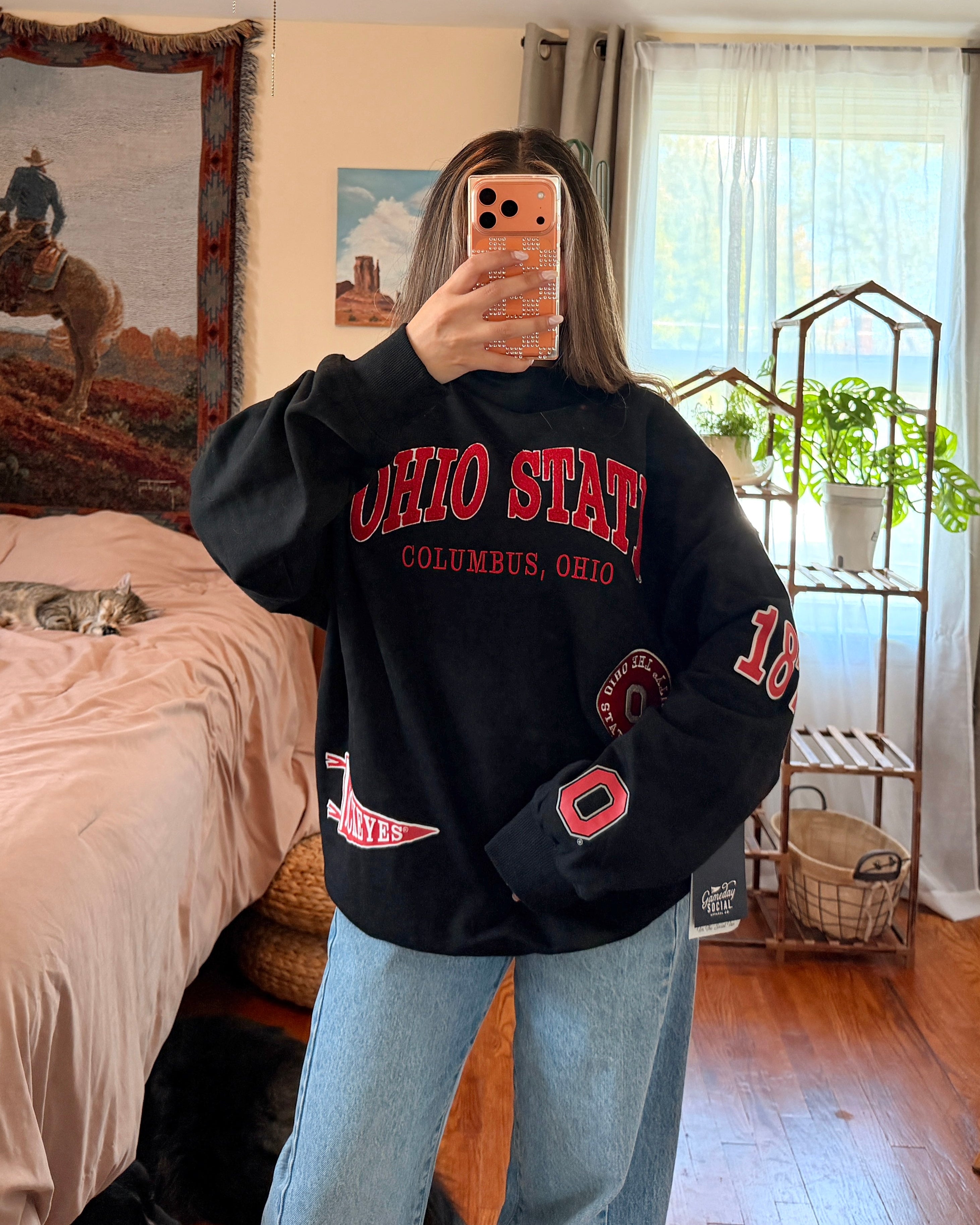 A person taking a mirror selfie in a bedroom, wearing a black Ohio State Buckeyes sweatshirt with red lettering and light blue jeans.