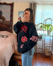A woman takes a mirror selfie in a bedroom, wearing a black sweatshirt with red collegiate-style lettering and light blue jeans. The room has a wooden floor, a bed with pink bedding, and a window with sheer curtains.