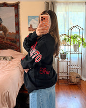 A woman takes a mirror selfie wearing a black sweatshirt with red lettering with light blue wide-leg jeans.