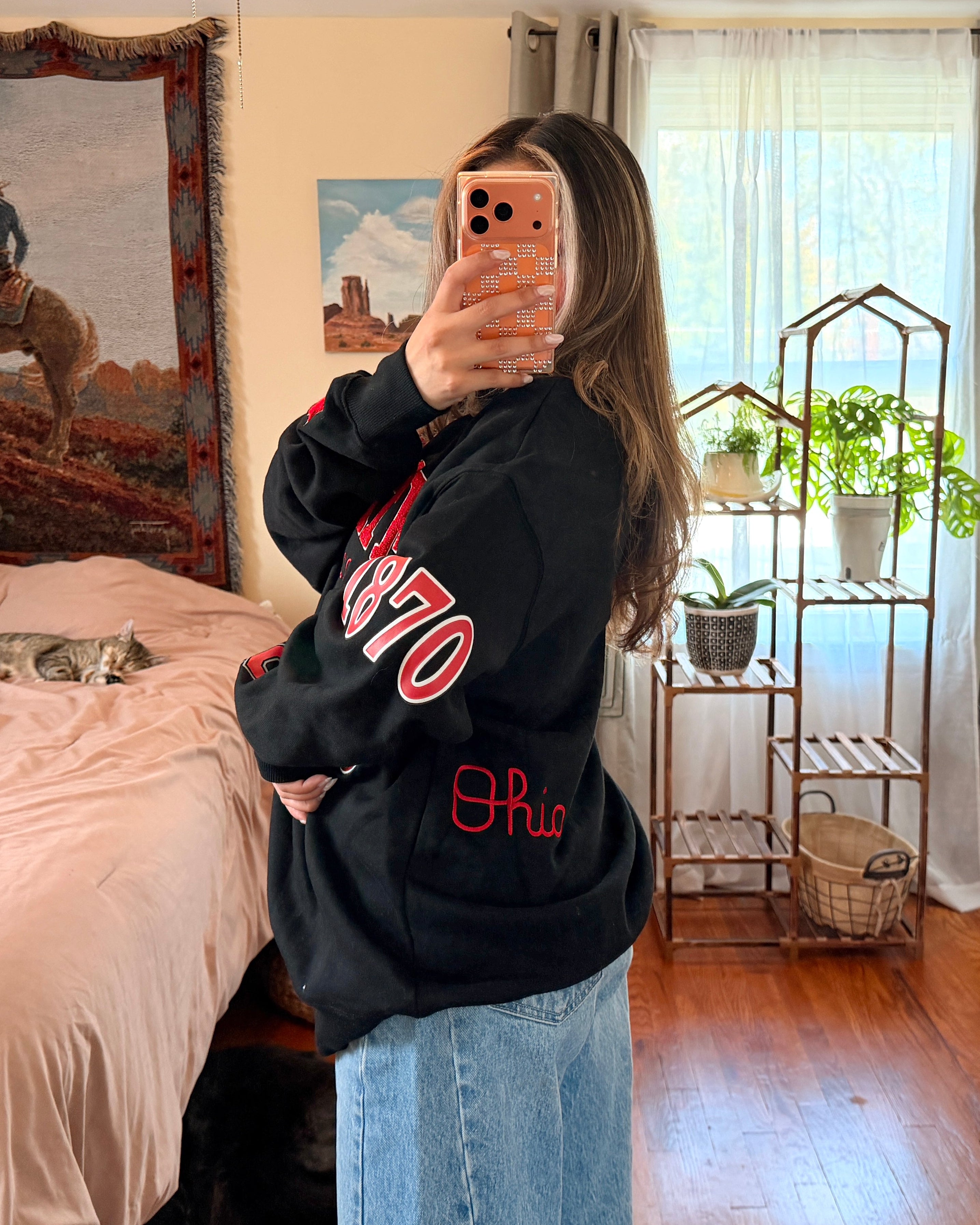 A woman takes a mirror selfie wearing a black sweatshirt with red lettering with light blue wide-leg jeans.