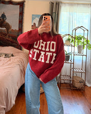 A woman takes a mirror selfie in a bedroom, wearing a red knit sweater with 'OHIO STATE' printed in white across the front, paired with light blue jeans.