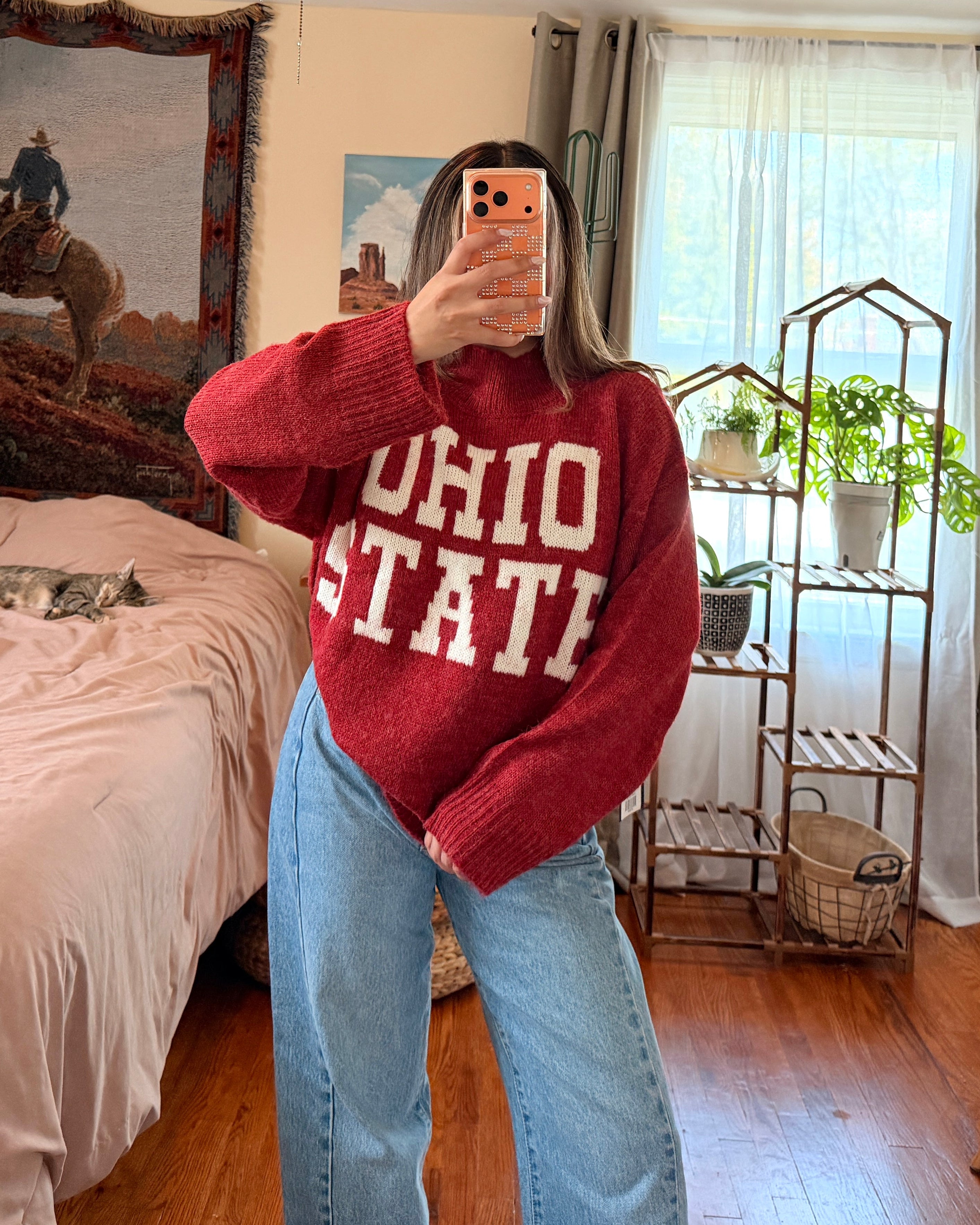 A woman takes a mirror selfie in a bedroom, wearing a red knit sweater with 'OHIO STATE' printed in white across the front, paired with light blue jeans.