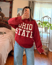 A woman takes a mirror selfie in a bedroom, wearing a red knit sweater with 'OHIO STATE' in white block letters and light blue jeans.
