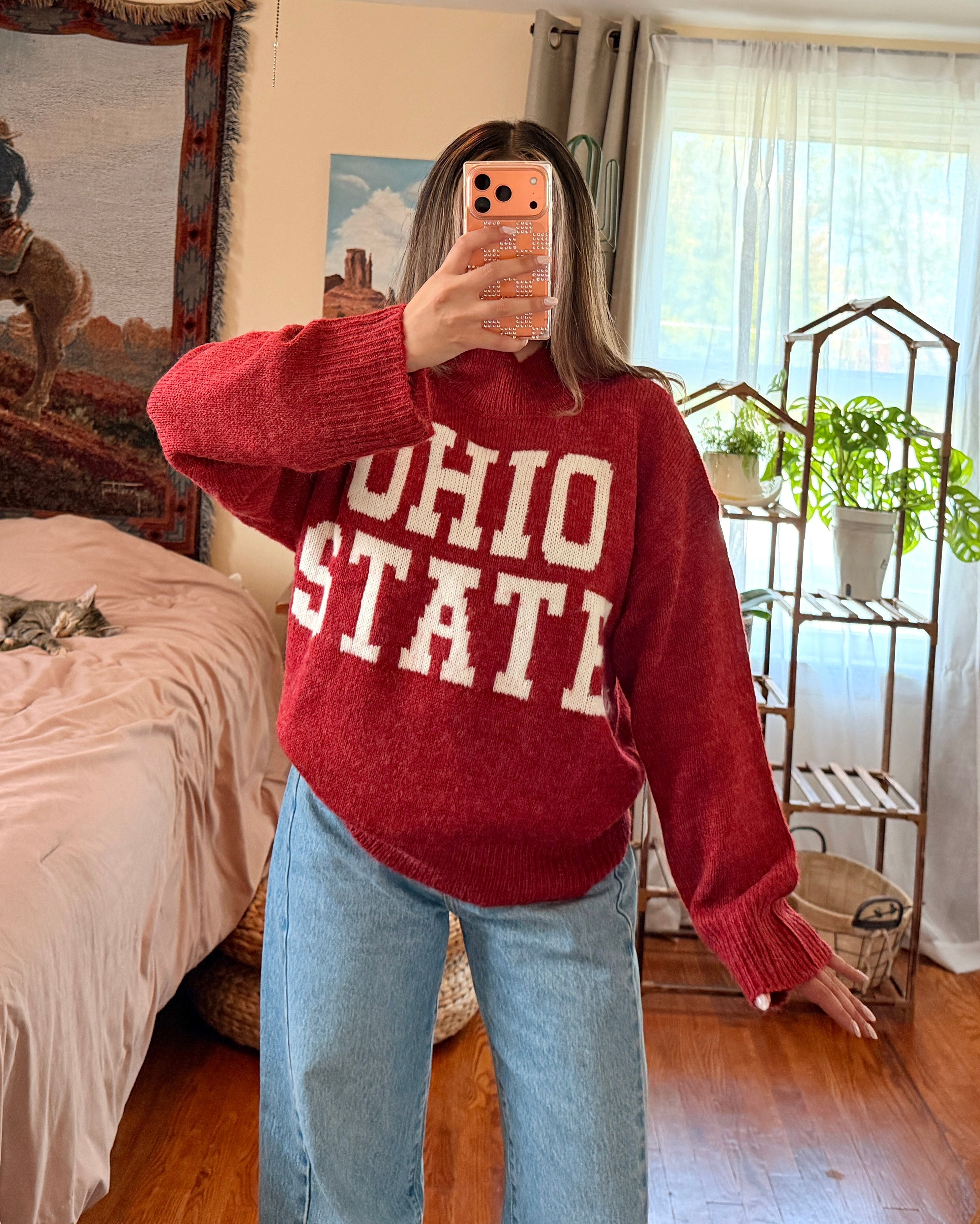 A woman takes a mirror selfie in a bedroom, wearing a red knit sweater with 'OHIO STATE' in white block letters and light blue jeans.
