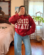 A person taking a mirror selfie in a bedroom, wearing a red knit sweater with 'OHIO STATE' printed in white across the front, paired with light blue wide-leg jeans.