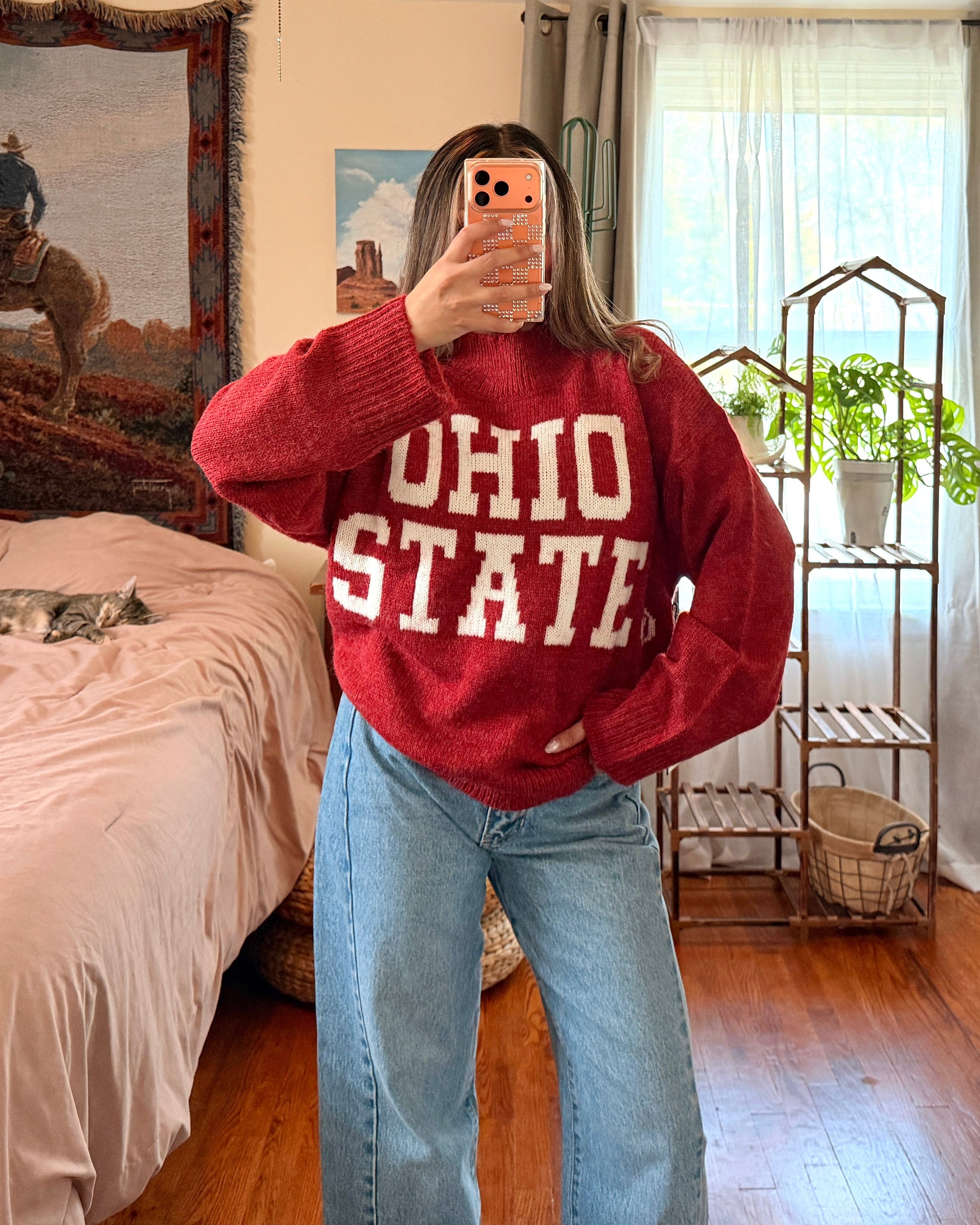 A person taking a mirror selfie in a bedroom, wearing a red knit sweater with 'OHIO STATE' printed in white across the front, paired with light blue wide-leg jeans.