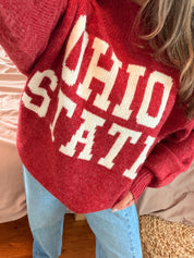 A close-up of a person wearing a red knit sweater with large white 'OHIO STATE' lettering, paired with light blue jeans in a bedroom setting.