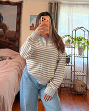 A woman takes a mirror selfie in a bedroom, wearing a striped button up cardigan knit sweater and light blue jeans.