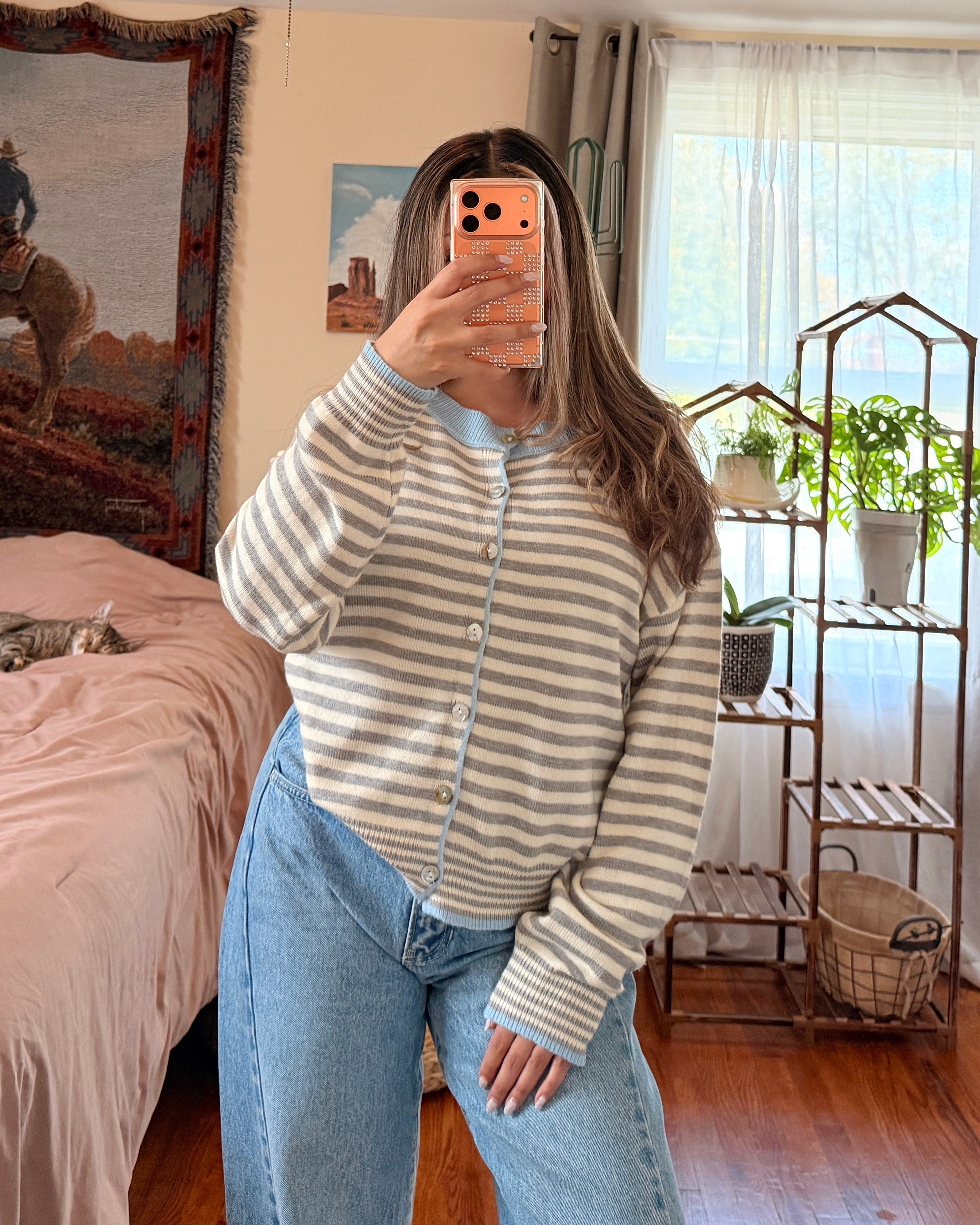 A woman takes a mirror selfie in a bedroom, wearing a striped button up cardigan knit sweater and light blue jeans.