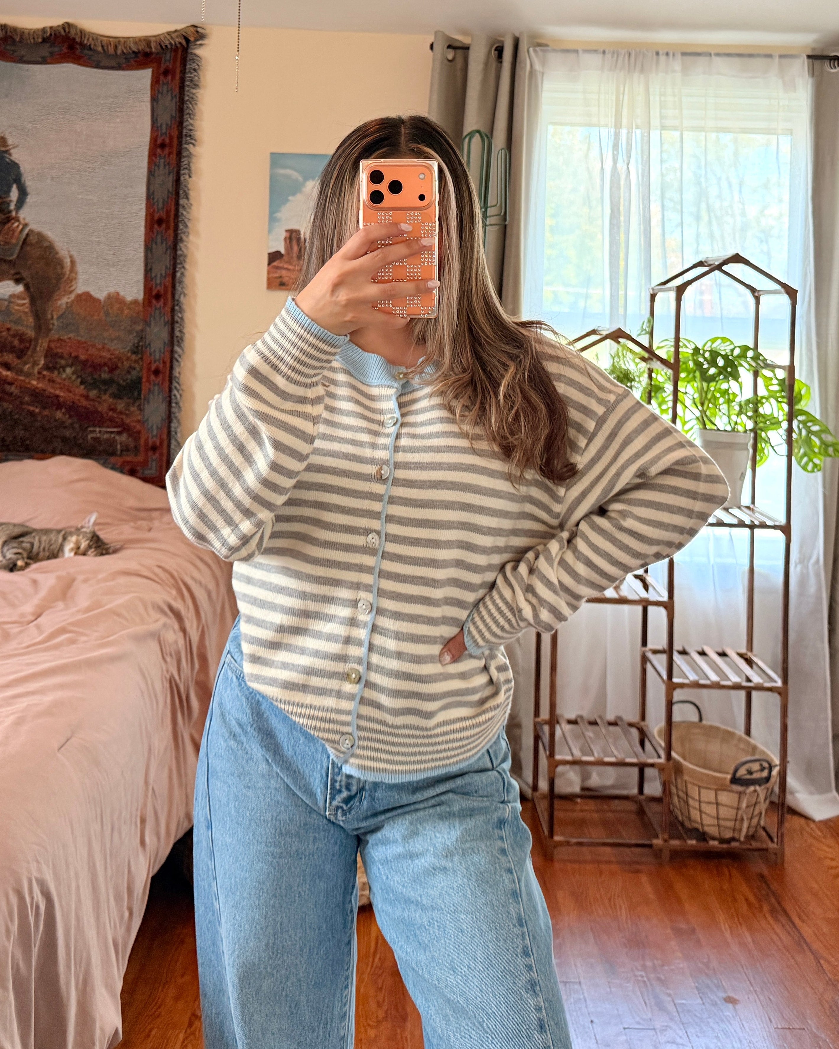 A woman takes a mirror selfie in a bedroom, wearing a striped button up cardigan knit sweater and light blue jeans.
