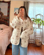 A woman takes a mirror selfie in a bedroom, wearing a cream-colored, textured crochet cardigan with brown floral patterns and blue jeans.