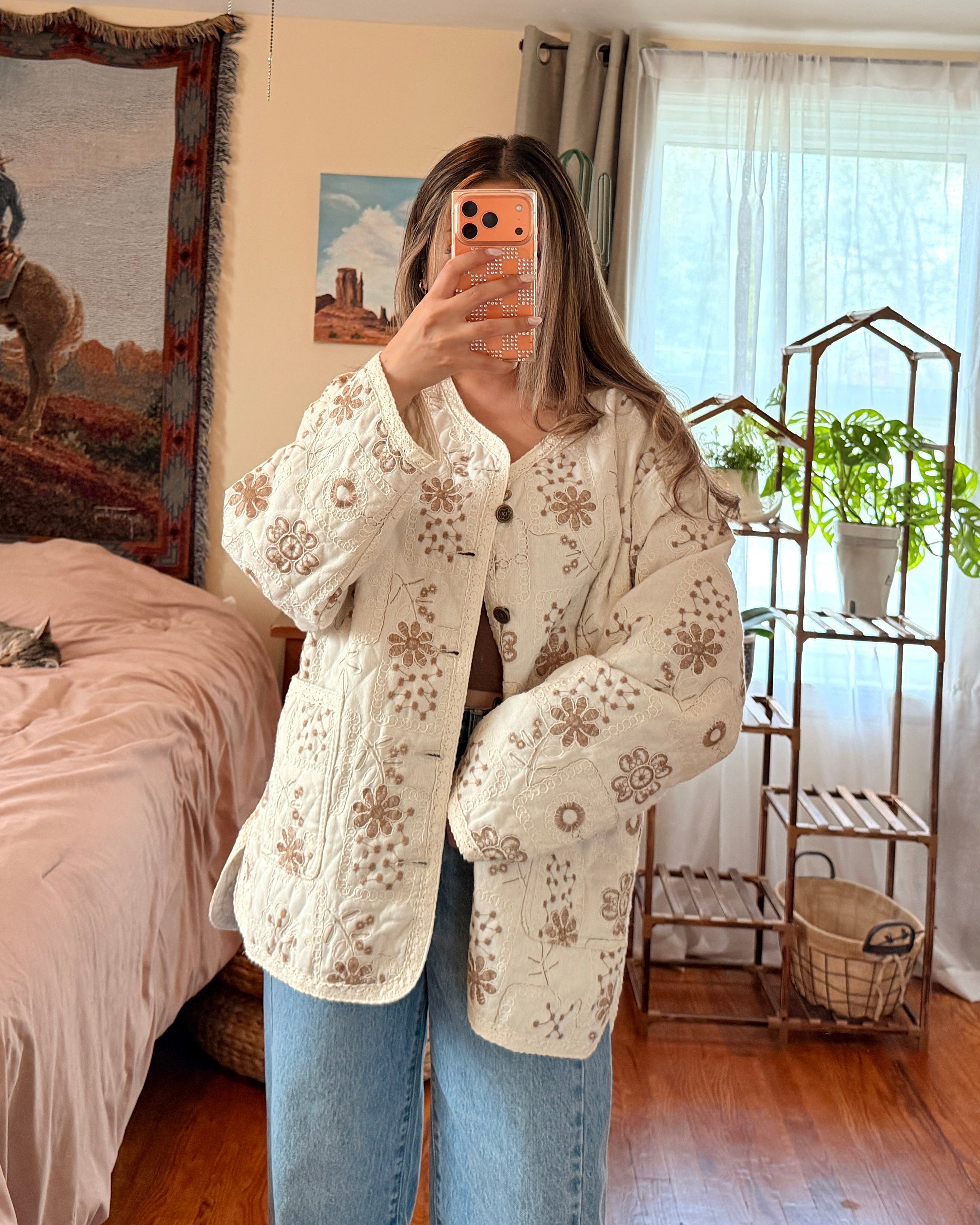 A woman takes a mirror selfie in a bedroom, wearing a cream-colored, textured crochet cardigan with brown floral patterns and blue jeans.