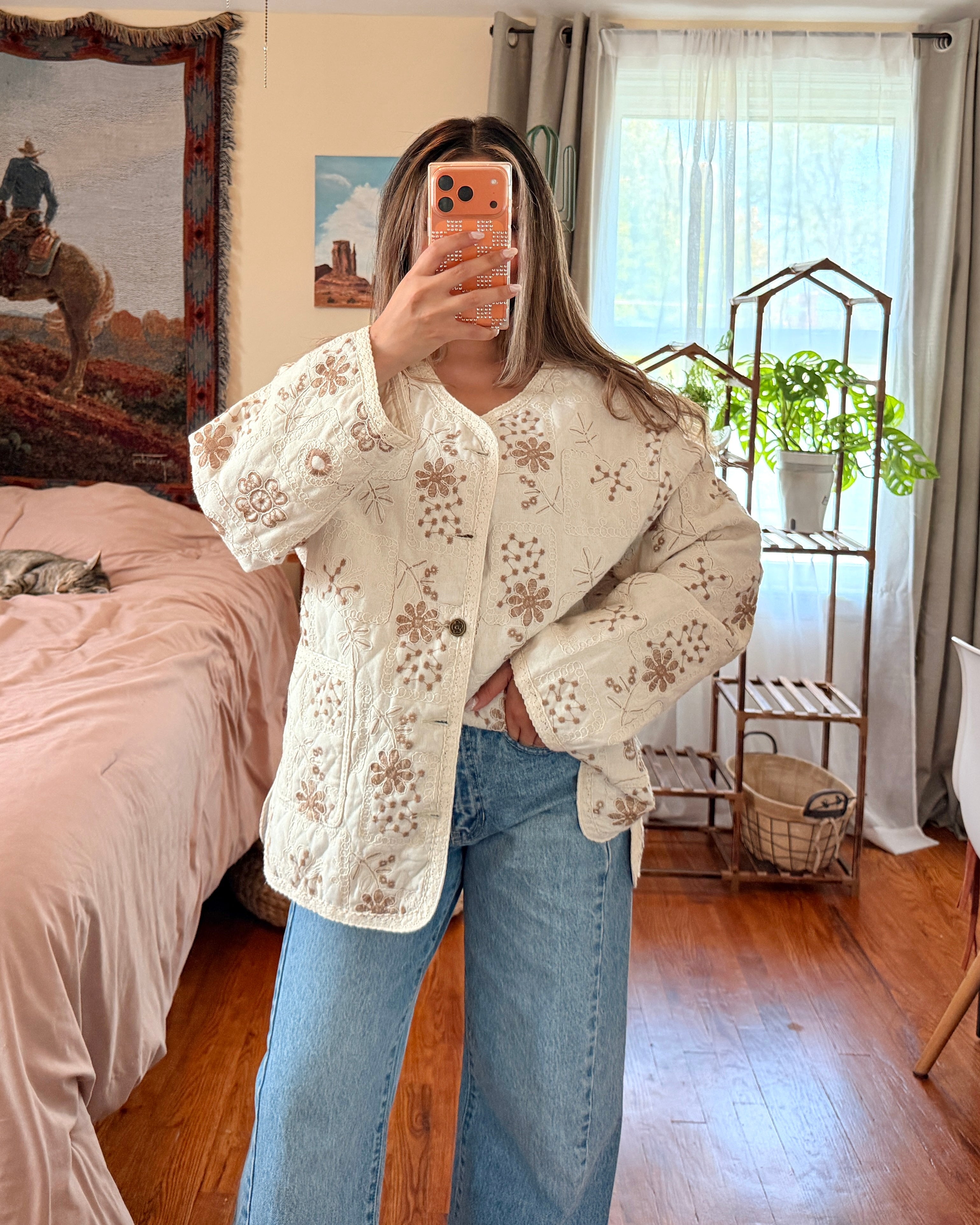 A woman takes a mirror selfie in a bedroom, wearing a cream-colored, button-front cardigan with intricate floral lace crochet detailing and blue jeans.