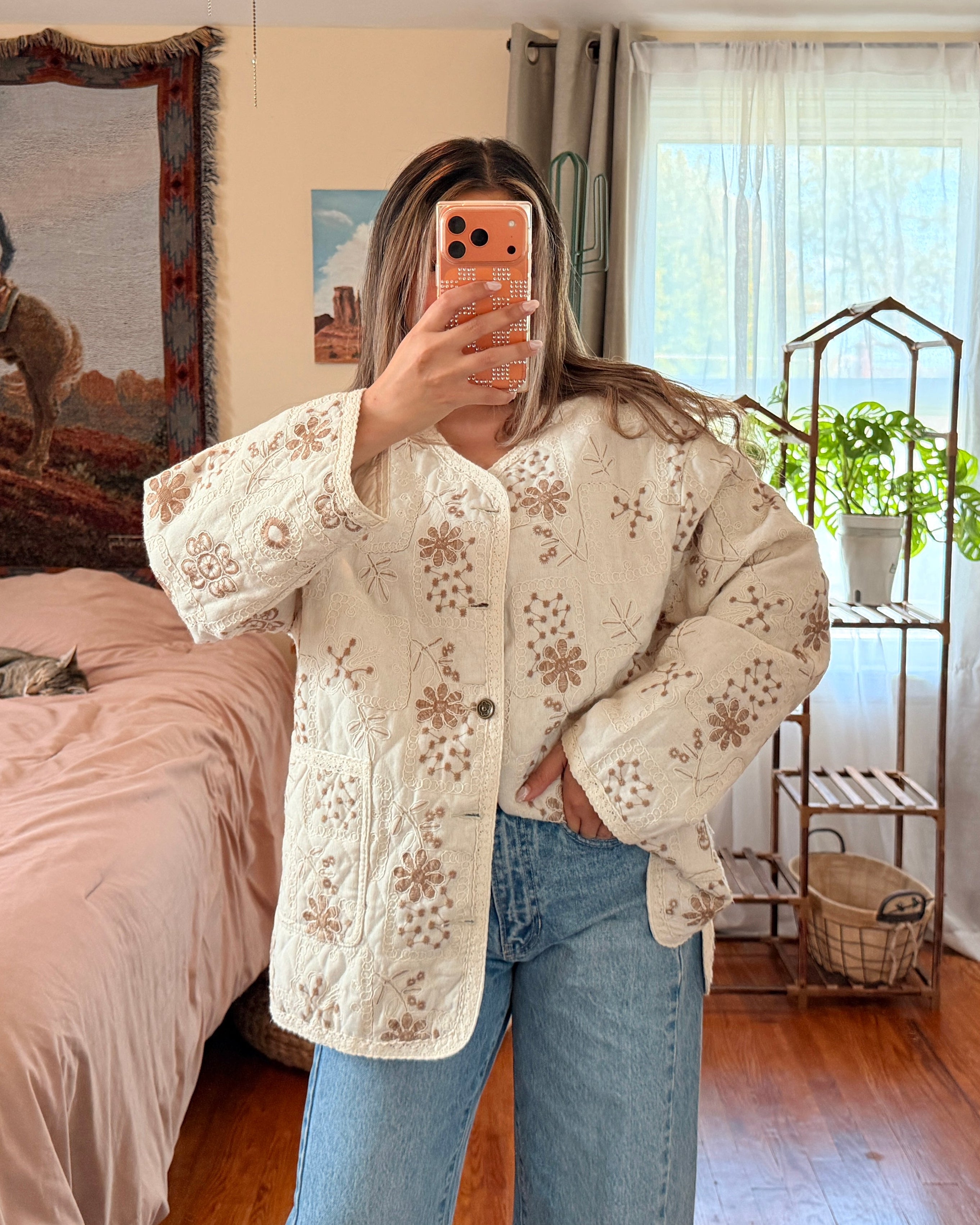 A woman takes a mirror selfie in a bedroom, wearing a cream-colored button-up shirt with brown floral embroidery and blue jeans.