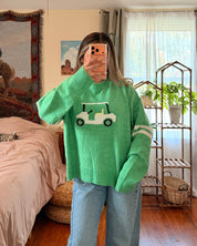 A person taking a mirror selfie in a cozy bedroom, wearing a bright green crewneck sweater with a white golf ball graphic on the chest and light blue jeans.