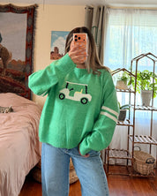 A woman takes a mirror selfie in a bedroom, wearing a bright green crewneck sweater with a white golf cart graphic on the front and light blue wide-leg jeans.