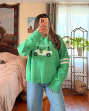 A woman takes a mirror selfie in a cozy bedroom, wearing a bright green crewneck sweater with a white and green golf cart graphic on the chest, paired with light blue wide-leg jeans.