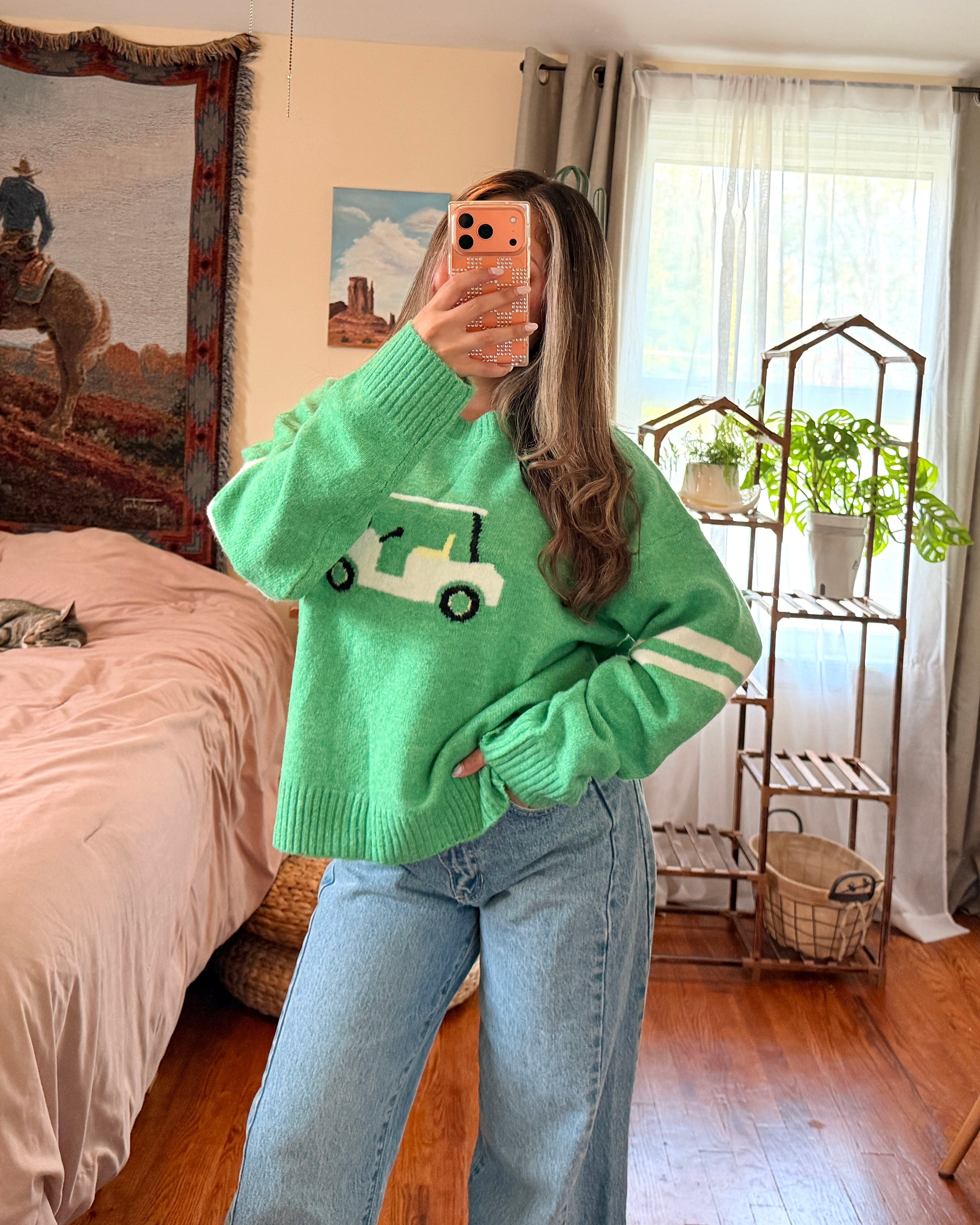 A woman takes a mirror selfie in a bedroom, wearing a bright green crewneck sweater with a white golf ball and club graphic on the chest, paired with light blue jeans.