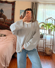 A woman takes a mirror selfie in a bedroom, wearing a striped long-sleeve shirt and light blue wide-leg jeans.