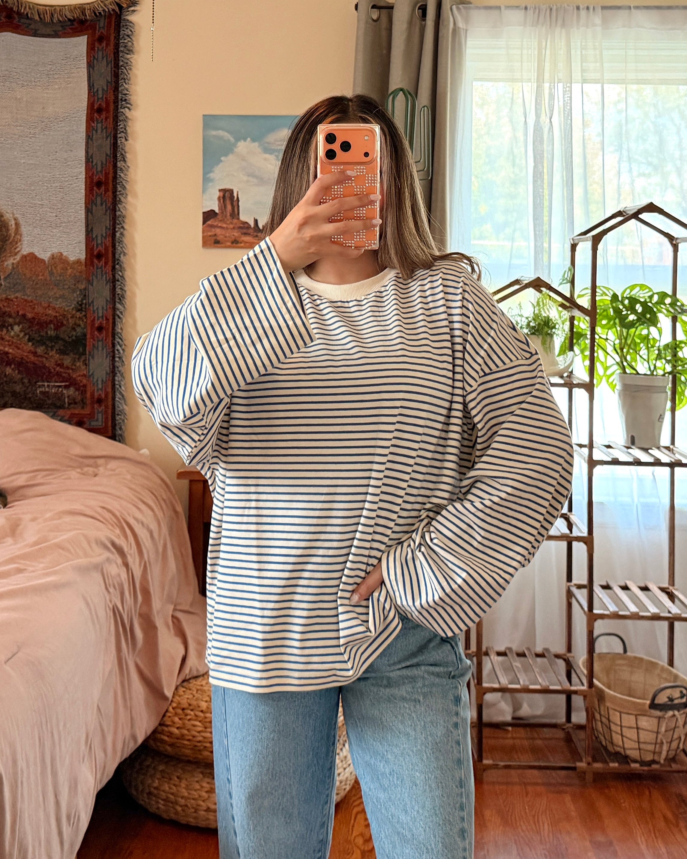 A woman takes a mirror selfie in a bedroom, wearing a striped long-sleeve shirt and light blue wide-leg jeans.