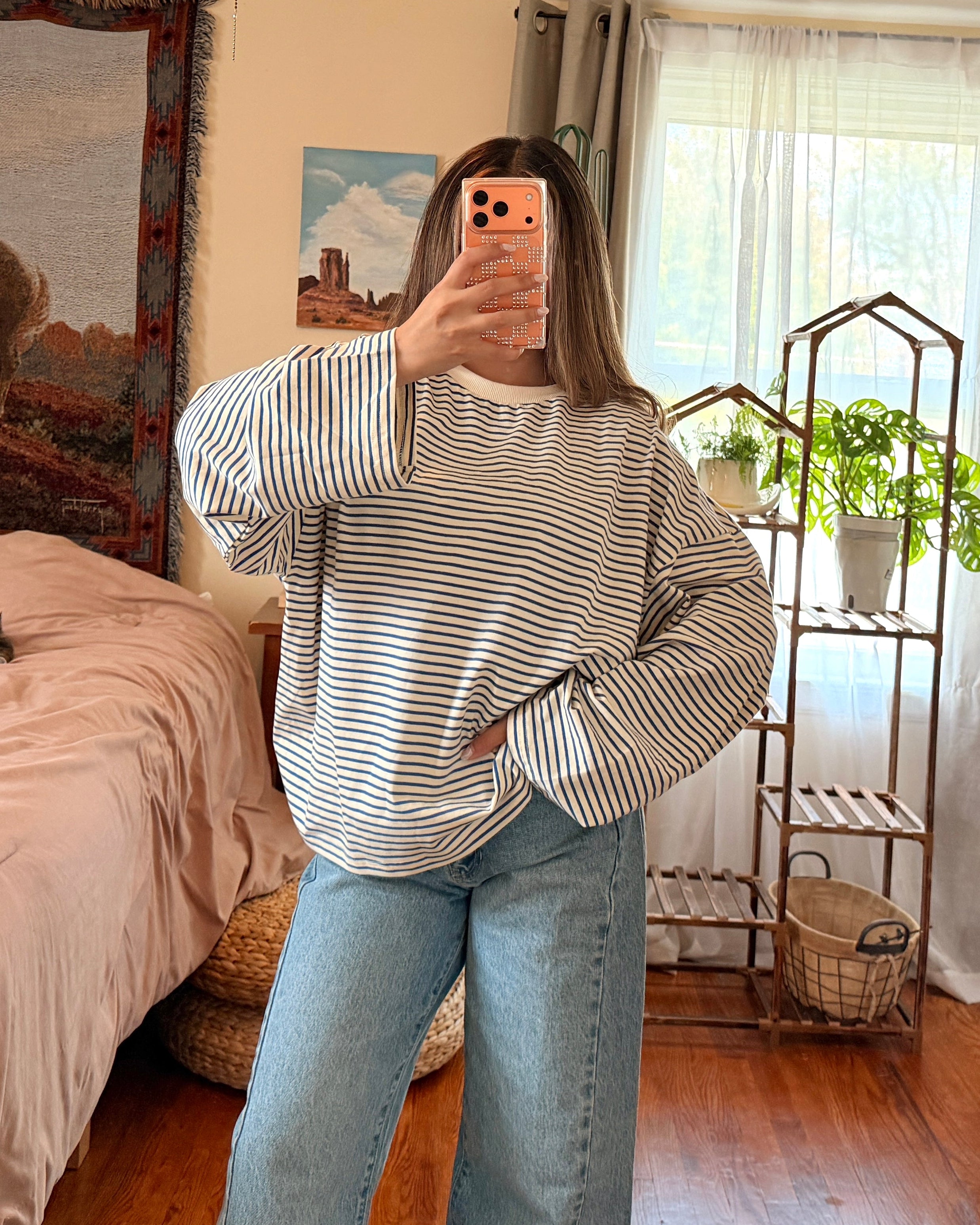 A woman takes a mirror selfie in a bedroom, wearing a striped long-sleeve shirt and light blue wide-leg jeans.