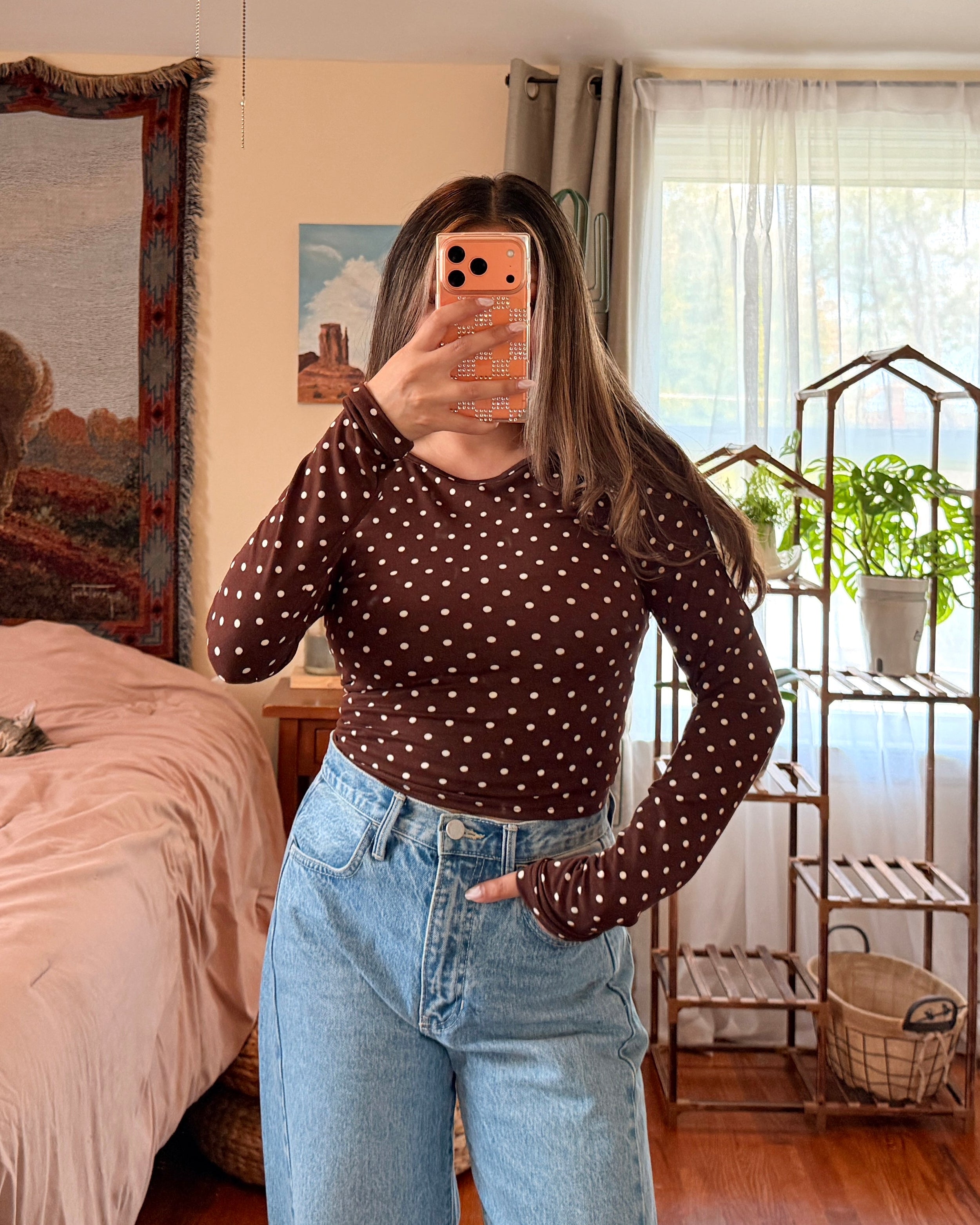 A woman takes a mirror selfie in a bedroom, wearing a dark brown long-sleeve crop top with white polka dots and light blue high-waisted wide-leg jeans.