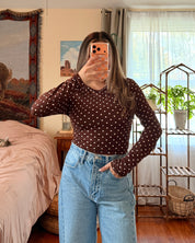 A woman takes a mirror selfie in a bedroom, wearing a dark brown long-sleeve top with white polka dots and light blue high-waisted wide-leg jeans.