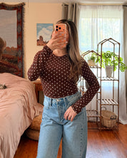 A woman takes a mirror selfie in a bedroom, wearing a dark brown long-sleeve top with small white polka dots and light blue high-waisted wide-leg jeans.