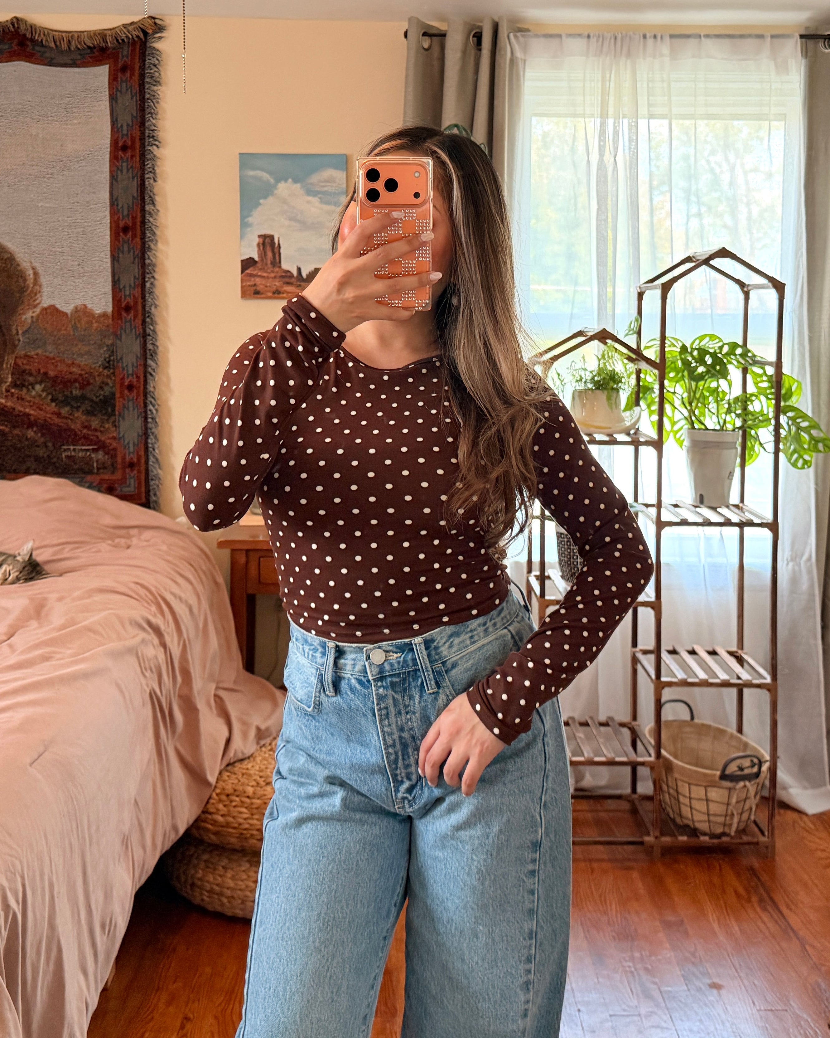 A woman takes a mirror selfie in a bedroom, wearing a dark brown long-sleeve top with small white polka dots and light blue high-waisted wide-leg jeans.