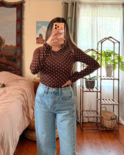A woman takes a mirror selfie in a bedroom, wearing a dark brown long-sleeve top with small white polka dots and light blue high-waisted wide-leg jeans.