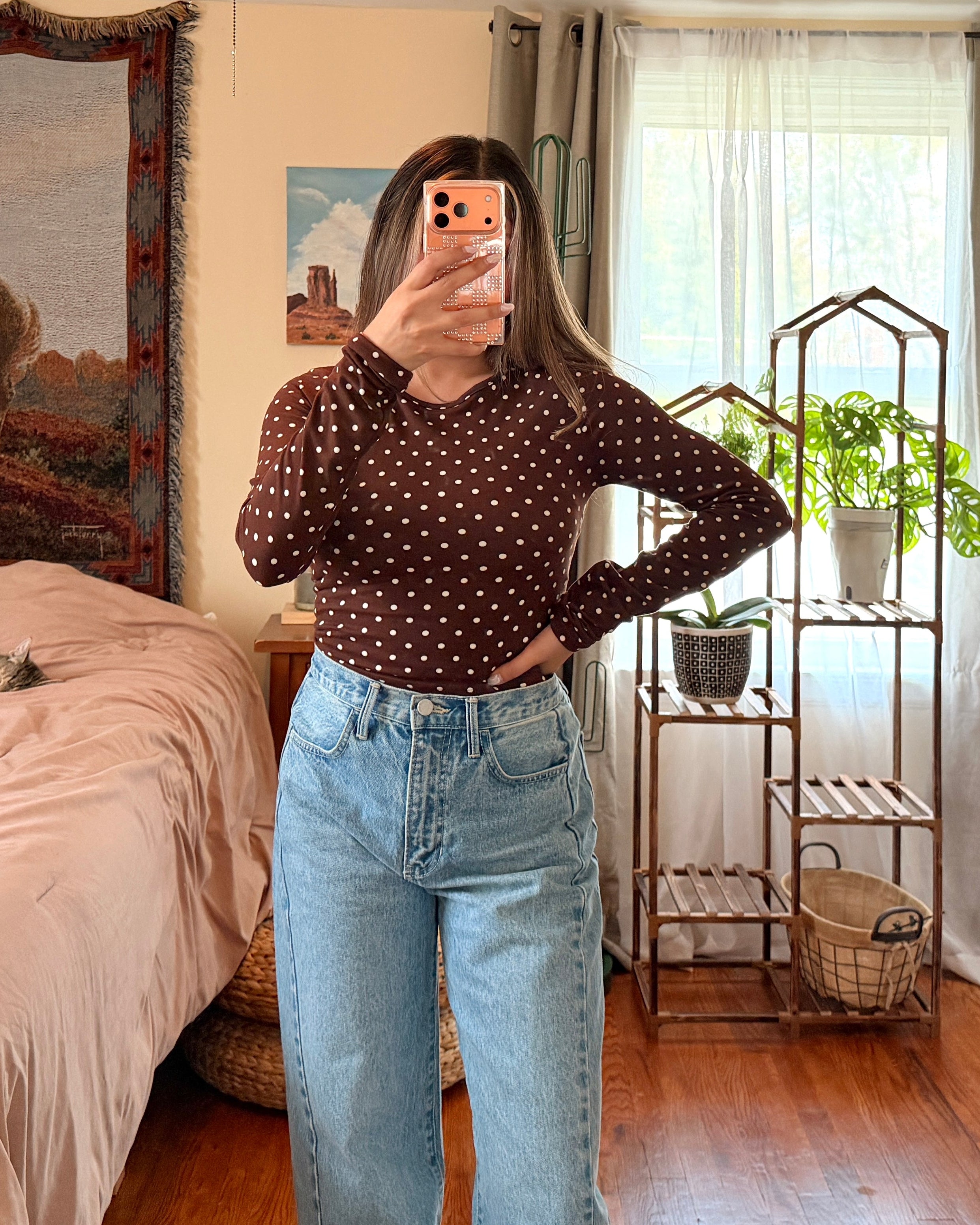 A woman takes a mirror selfie in a bedroom, wearing a dark brown long-sleeve top with small white polka dots and light blue high-waisted wide-leg jeans.