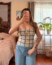 A woman takes a mirror selfie in a bedroom, wearing a beige and white plaid strapless tube top with light blue jeans.