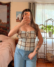 A woman takes a mirror selfie in a bedroom, wearing a beige and white plaid strapless tube top with light blue jeans.