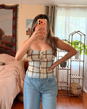 A woman takes a mirror selfie in a bedroom, wearing a beige and white plaid strapless tube top with light blue jeans.