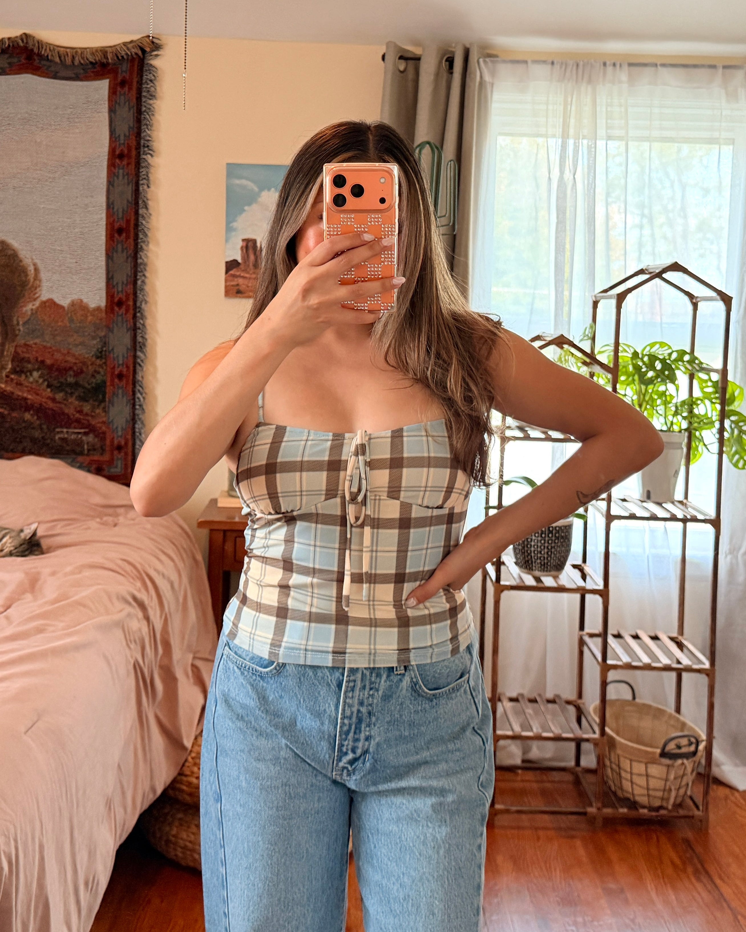 A woman takes a mirror selfie in a bedroom, wearing a beige and white plaid strapless tube top with light blue jeans.