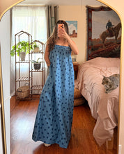 A woman takes a mirror selfie in a bedroom wearing a long blue maxi dress with a small black pattern. The room has a gold-framed arch mirror, wooden floors, a bed with pink bedding, and a cat lying on the bed.