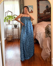 A woman takes a mirror selfie in a bedroom wearing a blue strapless maxi dress with a black polka dot pattern. The room features a gold-framed arched mirror, wooden floors, a bed with pink bedding