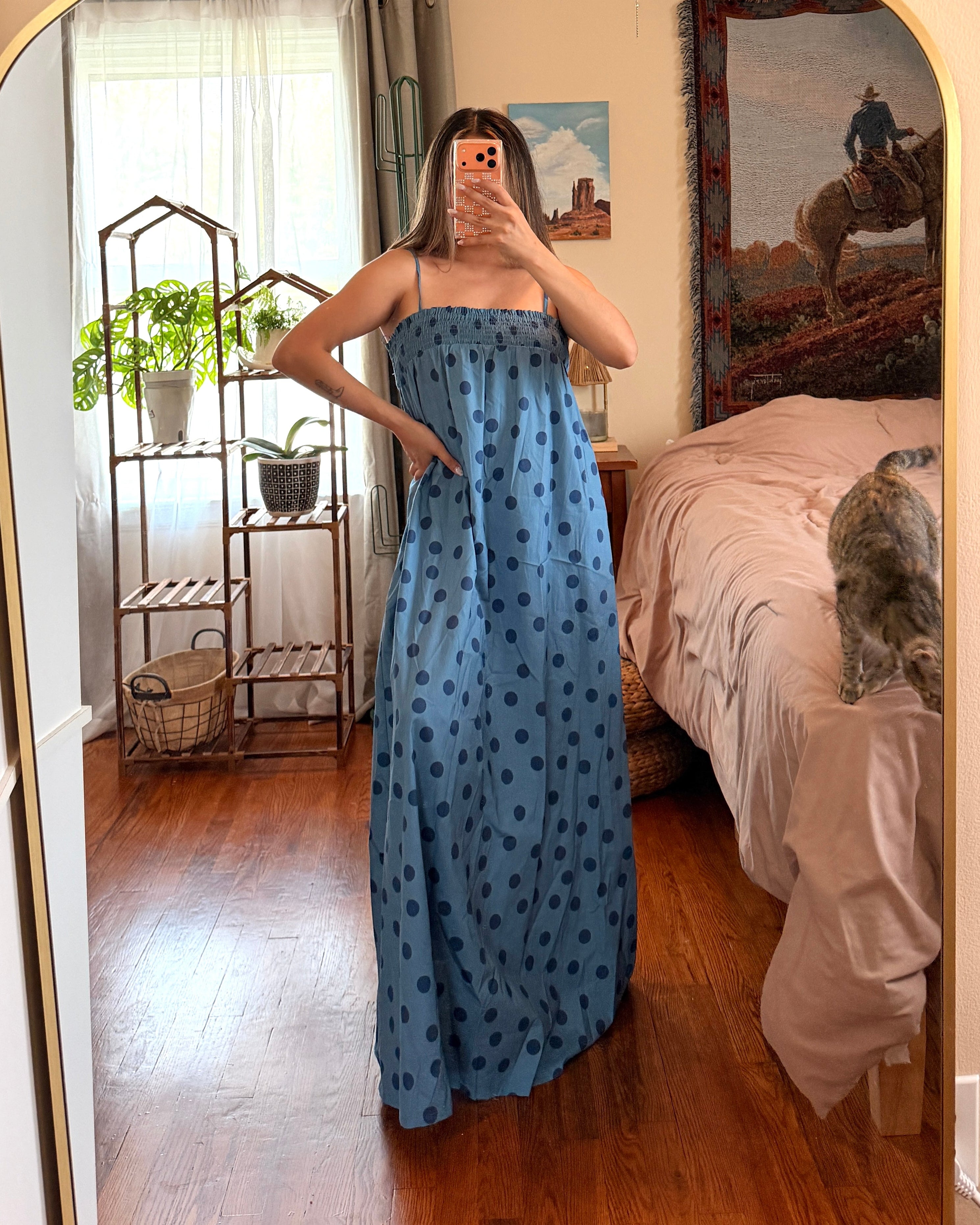 A woman takes a mirror selfie in a bedroom wearing a blue strapless maxi dress with a black polka dot pattern. The room features a gold-framed arched mirror, wooden floors, a bed with pink bedding