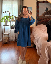 A woman takes a mirror selfie in a bedroom, wearing a blue smocked-embroidered long-sleeve dress with tie details at the cuffs. A black cat sits on the bed in the background, and a gold-framed archway surrounds the mirror.